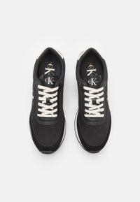 Zapatillas negras con cordones blancos, con una parte superior de textil suave, punta redondeada y acentos de suela blanca contrastantes. Logo visible en la lengüeta.