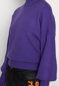 Maglione viola lavorato a maglia con collo alto, spalle scese e polsini a costine. Il design presenta una vestibilità comoda ed è abbinato a jeans scuri.