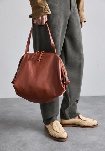 Brown leather handbag with a rounded shape, featuring a top handle, lacing detail, and a smooth texture. Worn with olive green pants.