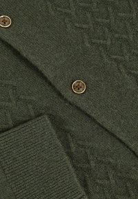 Dunkelgrüner Strickstoff mit geripptem Manschettenabschluss, goldgerahmtem Knopf und dezentem Rautenmuster.