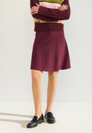 Personne debout, bras croisés, portant une jupe bordeaux, un pull rayé multicolore et des mocassins en cuir noir sur un fond uni.