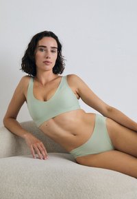 En kvinne med krøllete hår poserer på en teksturert sofa, iført en lys grønn sports-bh og matchende truse mot en nøytral bakgrunn.