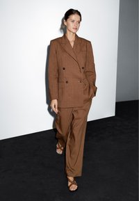 Brązouwy dwurzędowy garnitur z szerokimi spodniami i usztywnianą marynarką, wykonany z teksturowanej tkaniny. Modelka nosi sandały na obcasie na ciemnej podłodze.