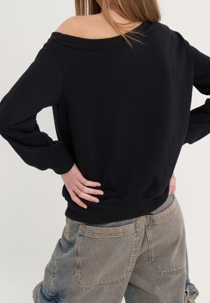 Personne portant un pull noir épaules dénudées et un jean gris délavé ample, les mains reposées sur le bas du dos, tournée de dos à la caméra.