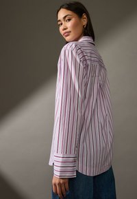 Camicia a righe bianca e rossa, con maniche lunghe, dettagli dorati sui bottoni e un fit rilassato. Indossata con pantaloni in denim blu.