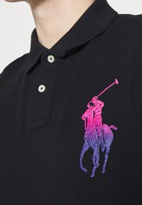Svart polotröja med texturerad stickning, med en gradient broderad logotyp av en häst och ryttare i rosa till lila nyanser. Tvåknappskrage.