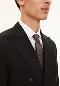 Close-up van een man in een zwart dubbelgebreid kostuum, met een witte overhemden en een gemêleerde bruine das, waarbij de nek en de onderkant van het gezicht zichtbaar zijn.