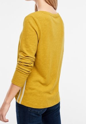 Vrouw die een mosterdgele lange mouwen shirt draagt met witte streepdetail aan de zijkant, gecombineerd met donkerblauwe spijkerbroek, gezien van achteren en zijkant.