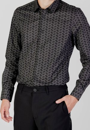 Mann trägt ein schwarzes, gemustertes Hemd mit Knöpfen und langen Ärmeln sowie eine einfarbige schwarze Hose, eine Hand in der Tasche, vor einem einfarbigen Hintergrund.