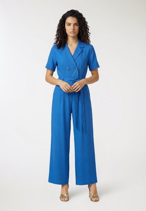 Donna con capelli ricci che indossa una tuta blu a maniche corte con pantaloni larghi e cintura in vita, in piedi con le mani intrecciate davanti.