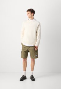 Pulover cu textură crem, pantaloni scurți cargo de culoare măslinie cu buzunare laterale, loafers negri și șosete albe. Modelul stă în fața unui fundal alb simplu.