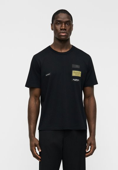 Μαύρο βαμβακερό T-shirt με πολλαπλά μπαλώματα με κείμενο. Σχέδιο με στρογγυλή λαιμόκοψη, κοντά μανίκια και ίσια ραφή. Φορεμένο με μαύρο παντελόνι.