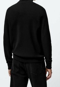 Personne portant un pull noir à manches longues et un jean noir, vue de dos sur un fond blanc uni.