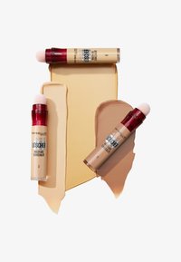 Correttori Maybelline in tre tonalità, con tubi cilindrici e tappi rossi, applicatori morbidi e campioni di colore beige cremoso su uno sfondo bianco.