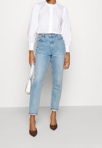 Femme portant un jean taille haute bleu clair, une blouse blanche à manches longues, tenant un petit sac à main blanc et des talons léopard marron.