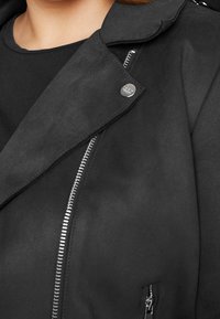 Giacca nera e strutturata con texture, caratterizzata da una zip diagonale, un bottone a pressione e una tasca laterale. Tessuto liscio e colletto strutturato con un design moderno.