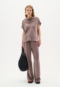 Top beige chiaro, morbido e ampio con scollatura a cisterna, abbinato a pantaloni larghi coordinati; tiene in mano una borsa a tracolla nera trapuntata. Sandali neri.