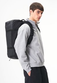 Schwarzer Rucksack mit mehreren Fächern und verstellbaren Trägern, getragen über einem hellgrauen Sweatshirt. Glattes Material, rechteckige Form, minimalistisches Design.