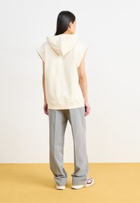 Cremfarbene ärmellose Kapuzenjacke aus weichem Material, kombiniert mit hellgrauen weiten Hose und weißen Sneakern mit roten Akzenten.