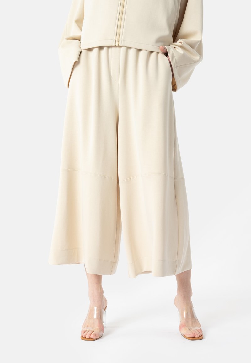 Personne portant un pantalon ample à jambes larges de couleur crème et des sandales à talons ouvertes et transparentes, debout les mains dans les poches sur fond blanc.