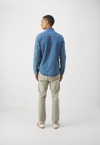 Camicia di denim blu medio con colletto e maniche lunghe, abbinata a pantaloni beige chiaro e sneaker bianche con accenti rossi.