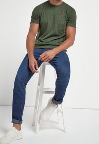 Grøn kortærmet t-shirt med et lille rødt logo, parret med mørkeblå jeans og hvide sneakers, siddende på en hvid skammel mod en ensfarvet baggrund.