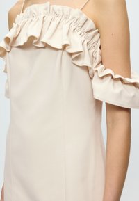 Εκρού αμάνικο φόρεμα με σούρες και βολάν κατά μήκος του ντεκολτέ και ανοίγματα στα μανίκια, φορεμένο από άτομο με ανοιχτό χρώμα επιδερμίδας.