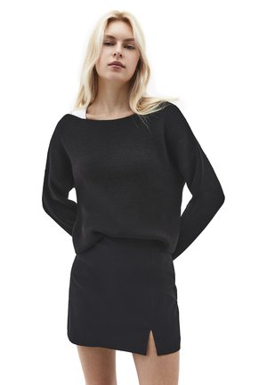 Blond kvinde iført en sort striktrøje over en hvid top og en sort mininederdel med en slids foran, stående med hænderne bag ryggen.