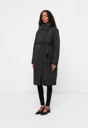 Schwarzer gepolsterter Mantel mit hohem Kragen, tailliertem Gürtel und langen Ärmeln, kombiniert mit schwarzen Leggings und Schuhen. Strukturiertes Material.