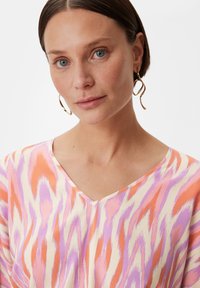 V-hals top van lichtgewicht stof met een abstract patroon in roze, oranje en lavendel; model met oorringen op een effen achtergrond.