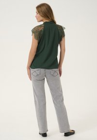 Blusa verde con maniche a cappuccio in pizzo, tessuto testurizzato, abbinata a jeans a gamba larga grigi chiari con dettagli delle tasche cuciti e scarpe nere.