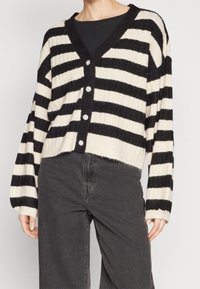 Czarno-kremowy sweter na guziki w paski z dekoltem w szpic i prążkowaną fakturą, zestawiony z ciemnoszarymi spodniami.