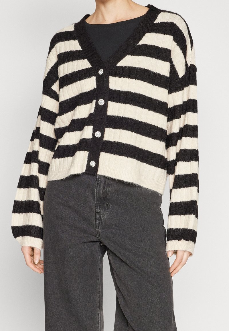 Czarno-kremowy sweter na guziki w paski z dekoltem w szpic i prążkowaną fakturą, zestawiony z ciemnoszarymi spodniami.