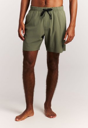 Man met olijfgroene zwembroek met een zwarte trekkoord, staand op blote voeten tegen een eenvoudige lichte achtergrond.