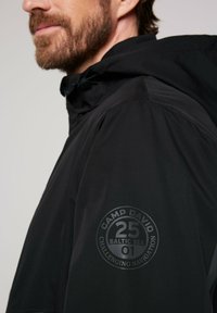 Giacca nera impermeabile con cappuccio. Presenta un logo rotondo sulla manica, che riporta la scritta "Camp David 25 Baltic Sea 01" con una texture liscia.