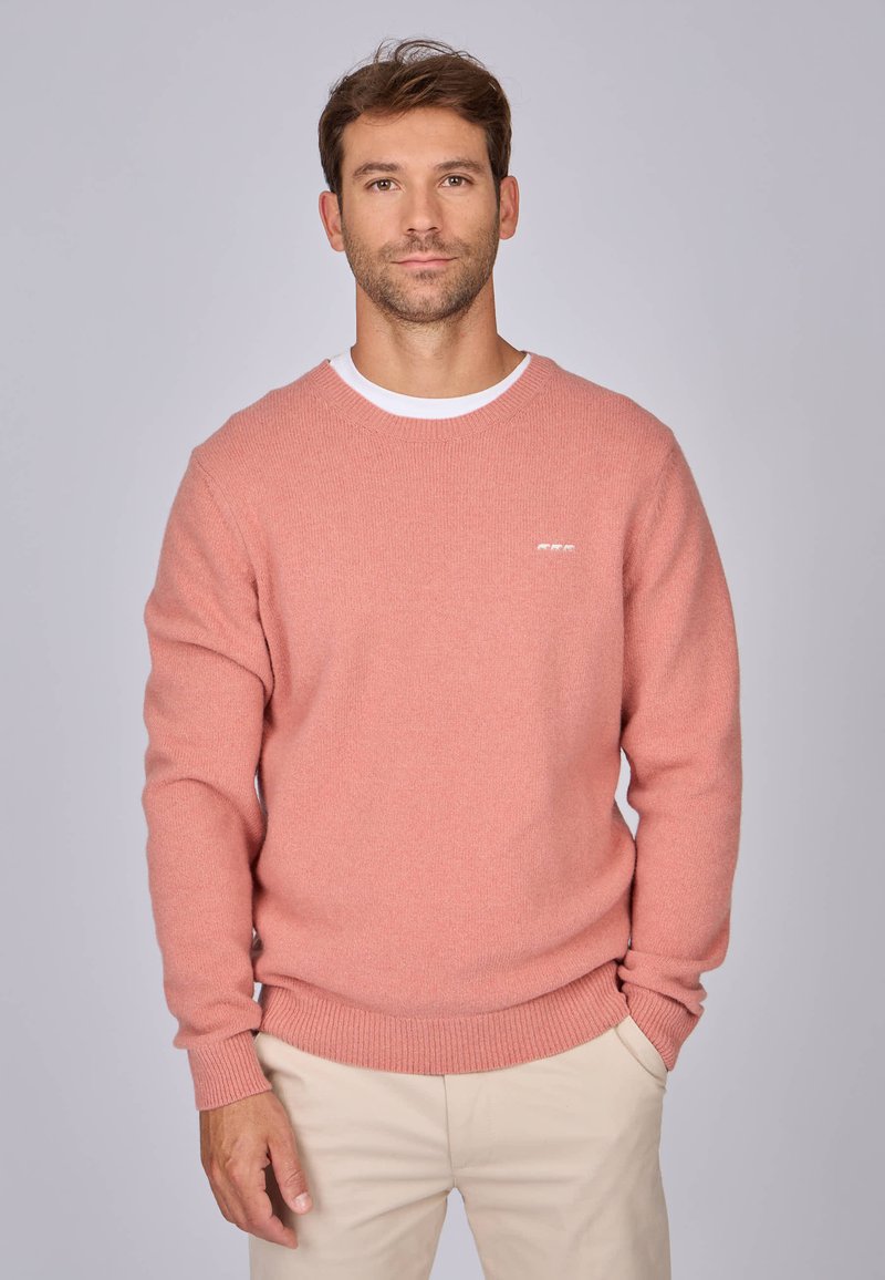 Pull en tricot rose avec des manches longues, un col rond et des poignets côtelés. Petit détail logo blanc sur la poitrine. Stylé par-dessus une chemise blanche.