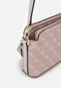 Helles rosa Umhängetasche mit weißem Logo-Muster, goldener Reißverschluss und beigem Riemen auf weißem Hintergrund.