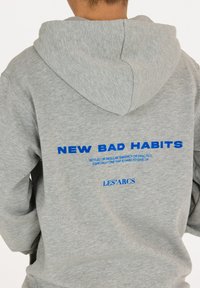 Grijze hoodie met een kangoeroezak en een trekkoord kap. Voorzien van blauwe tekst op de achterkant: "NEW BAD HABITS" en "LES'ARCS." Zachte textuur.