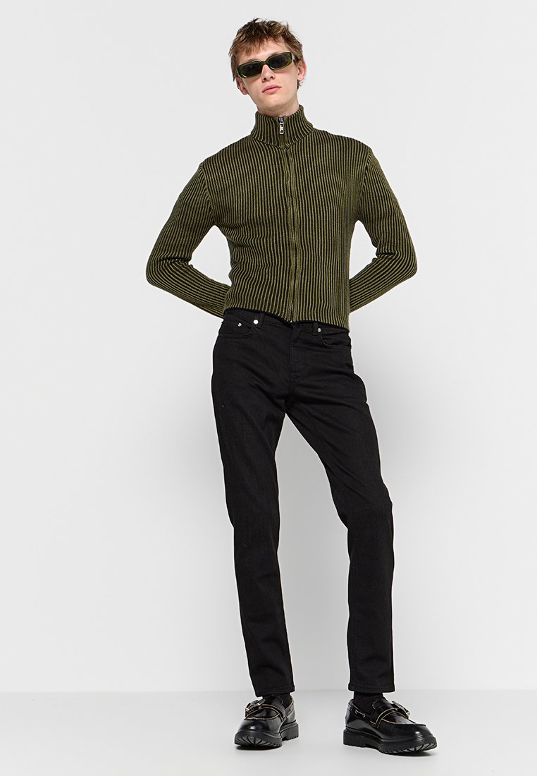 Olivgrüner, gerippter Reißverschluss-Pullover mit hohem Kragen; kombiniert mit schwarzen Hosen und klobigen schwarzen Schuhen. Das Model trägt dunkle Sonnenbrillen.