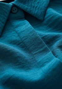 Teal linnen shirt met een gestructureerde kraag, een enkele knop bij de hals en zichtbare stikdetails. Zachte textuur en subtiele glans.