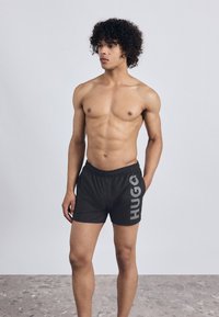 Melni peldbikses ar elastīgu jostasvietu ir ar lielu pelēku "HUGO" logo sānos. Audums izskatās viegls un gluds.