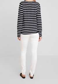 Maglia a maniche lunghe a righe navy e bianche con scollo rotondo, abbinata a jeans bianchi slim-fit e sandali neri. Vista posteriore mostrata.