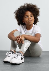 Criança vestindo uma t-shirt branca e leggings cinzas com um grande logótipo da Nike, combinadas com ténis Nike brancos e pretos, sentada sobre uma superfície cinza.