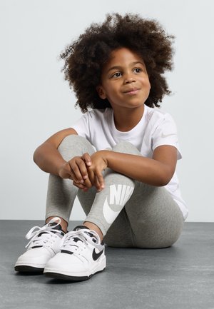 Niño vistiendo una camiseta blanca y leggings grises con un gran logo de Nike, combinado con zapatillas Nike blancas y negras, sentado sobre una superficie gris.