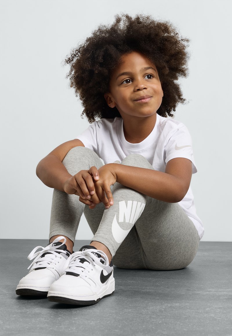 Criança vestindo uma t-shirt branca e leggings cinzas com um grande logótipo da Nike, combinadas com ténis Nike brancos e pretos, sentada sobre uma superfície cinza.