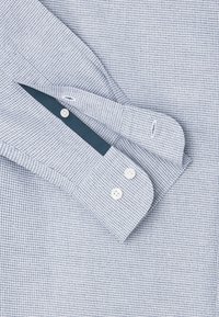Camicia a quadri blu con tessuto testurizzato, bottoni bianchi e un polsino interno a contrasto di colore blu scuro.