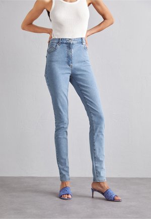 Jean taille haute bleu clair, coupe ajustée, en denim. Assortis à des talons ouverts à motifs bleus. Le haut du modèle est un débardeur côtelé blanc.