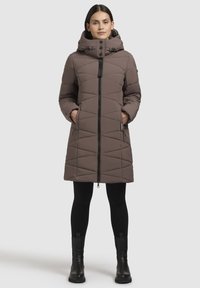 Bruine gewatteerde pufferjas met een hoge kraag en ritssluiting. Bevat zijzakken, een capuchon en is gecombineerd met zwarte leggings en laarzen.