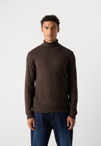 Brah z pulover s turtle neck iz mehkega, teksturiranega pletenega materiala. Ima dolge rokave in rebrast ovratnik, v kombinaciji z modrimi kavbojkami.