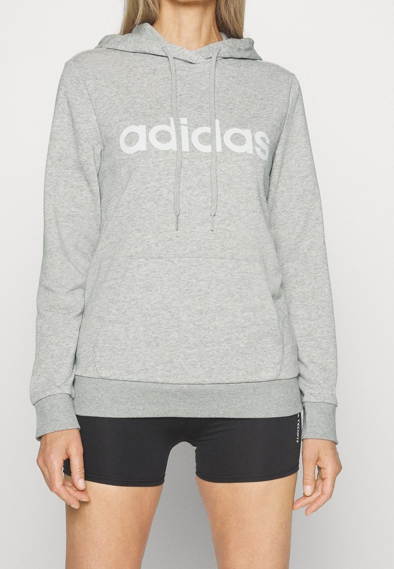 Personne portant un sweat à capuche gris clair avec le texte « adiclas » et un short de sport noir moulant sur un fond clair uni.